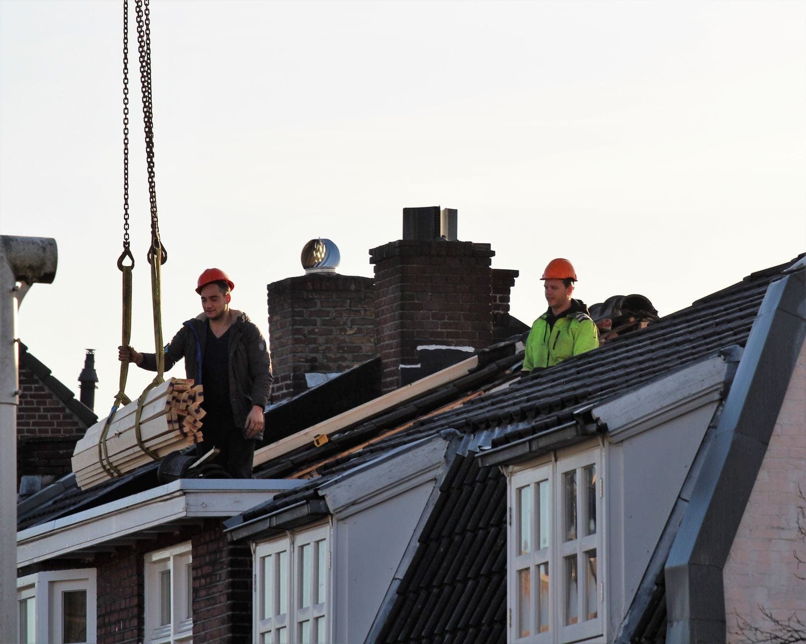 Roofing crew on a dark shingle roof lifting materials with a crane hoist