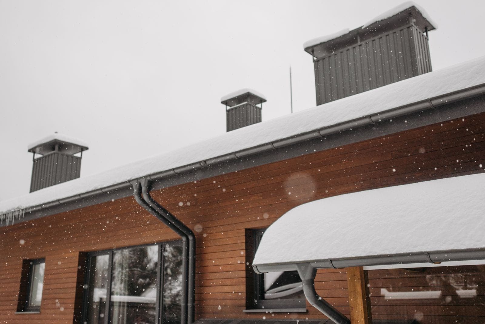 Snow-covered roof eave with gutter and downspout on a wood-sided building