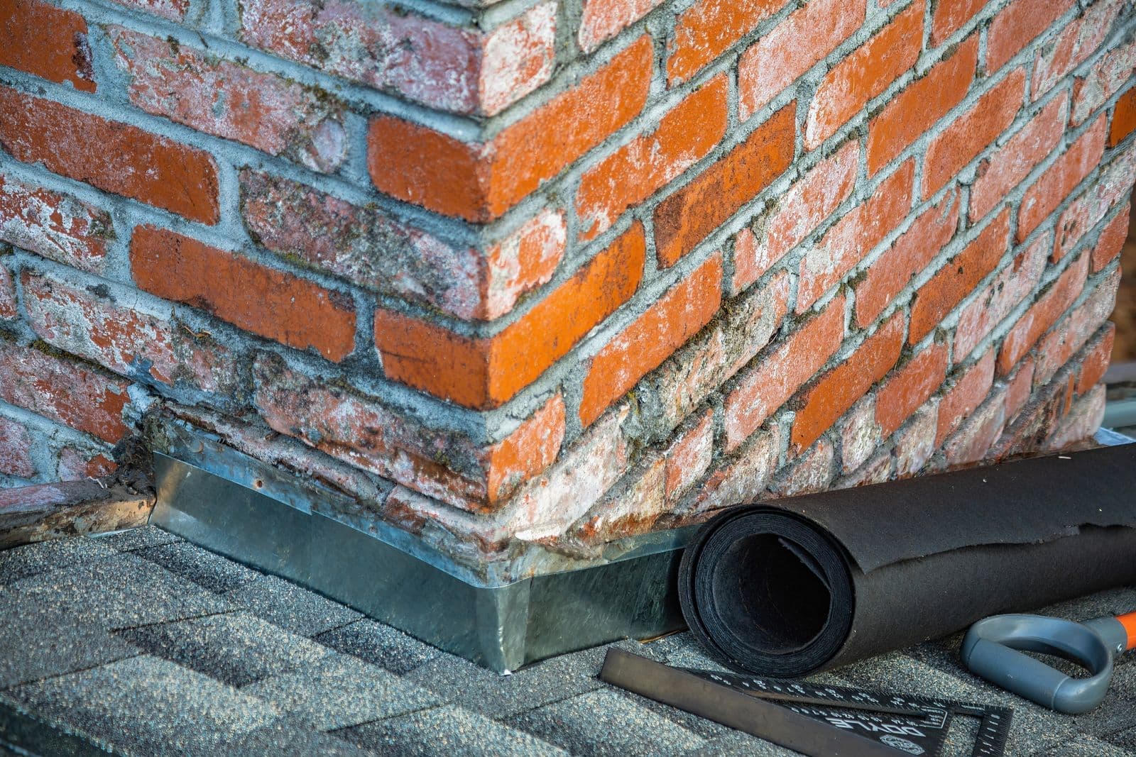 Chimney base with metal flashing and a roll of underlayment resting on asphalt shingles